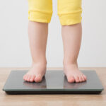 Baby,Girl,With,Barefoot,Standing,On,Weight,Scales,On,Wooden